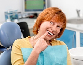 Older woman in dental chair pointing to her smile after dental implant retained denture placement