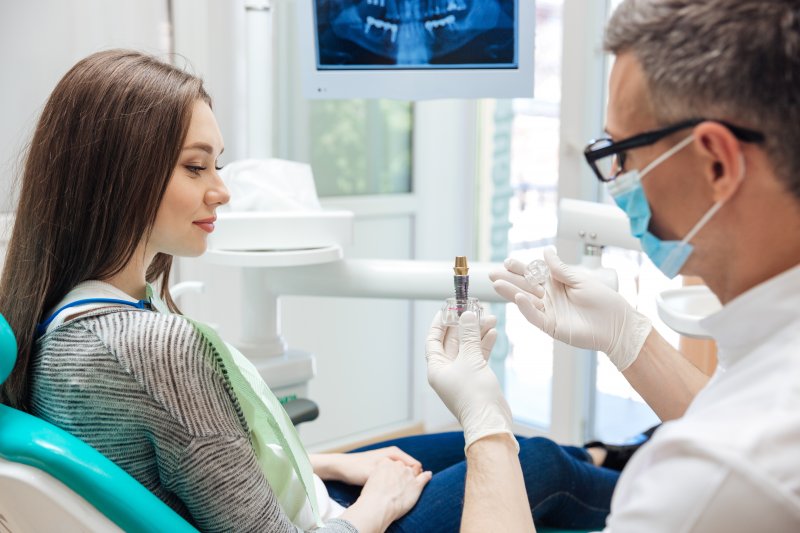 Masked dentist holding dental implant model with glover hand gesturing with his other hand in front of female patient in dental chair
